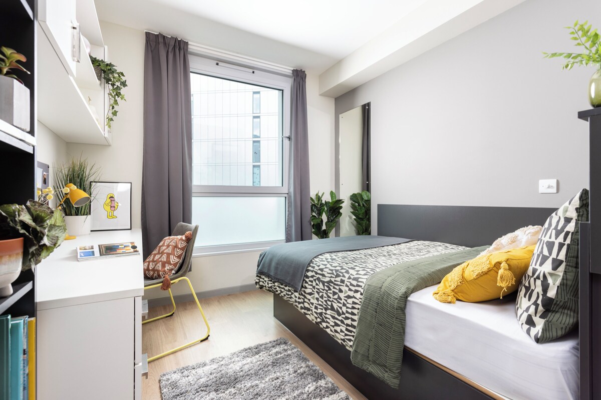 A modern bedroom with a bed, desk, chair, and large window.
