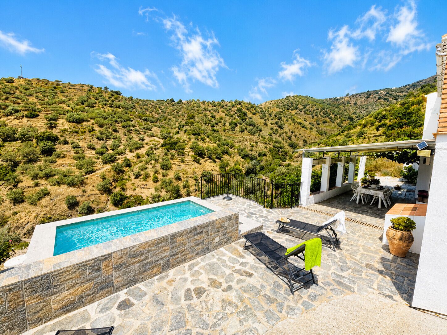 Ein Pool mit Steinfliesen, Liegestühlen, einem Tisch und einer Pergola mit Solarmodul.