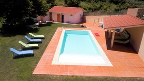 A rectangular swimming pool with a tiled deck, surrounded by grass and a small building with a red roof.
