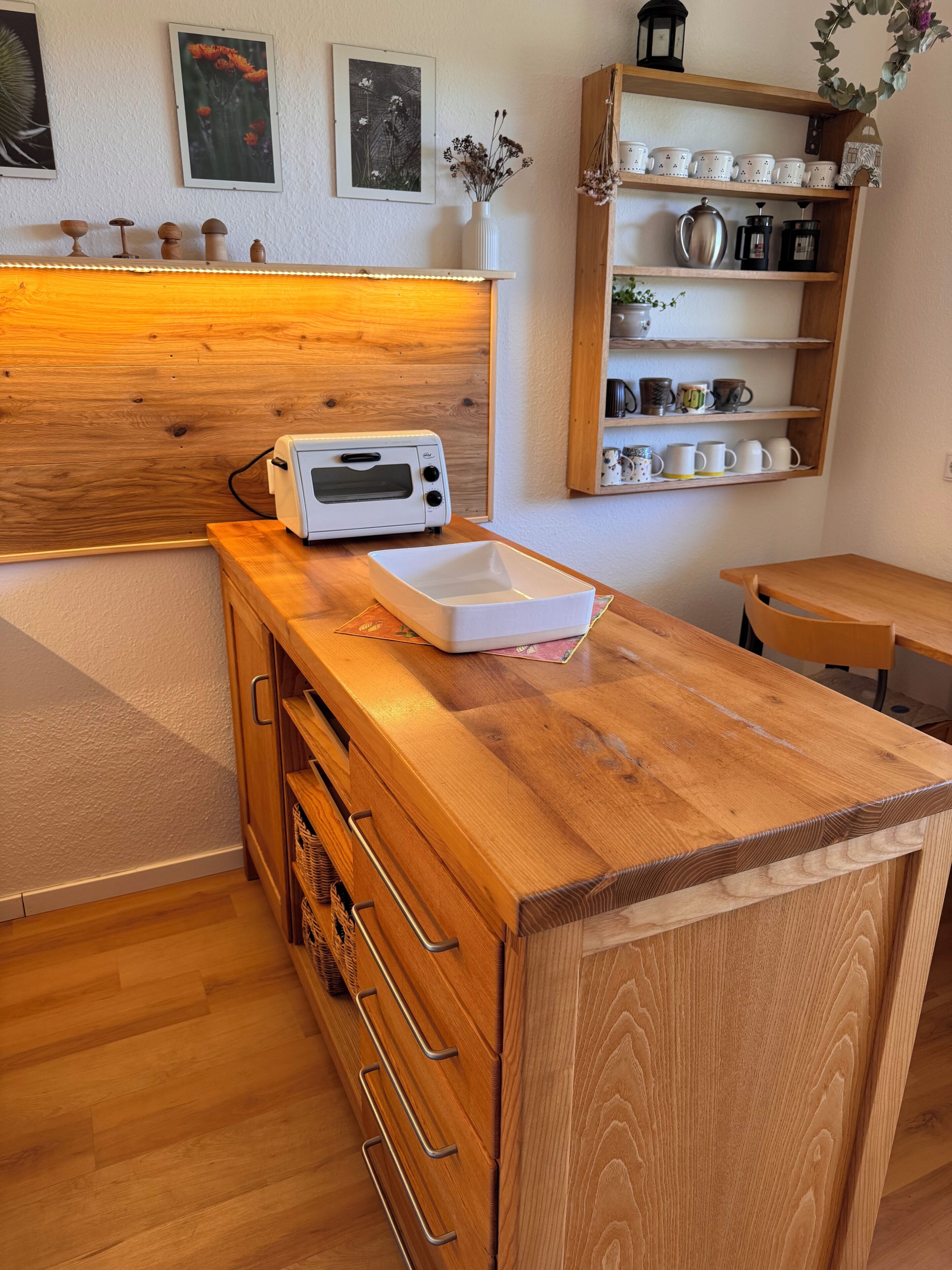 A kitchen with a wooden countertop, a toaster, a white vase with dried flowers, and a shelf with mugs and a teapot.