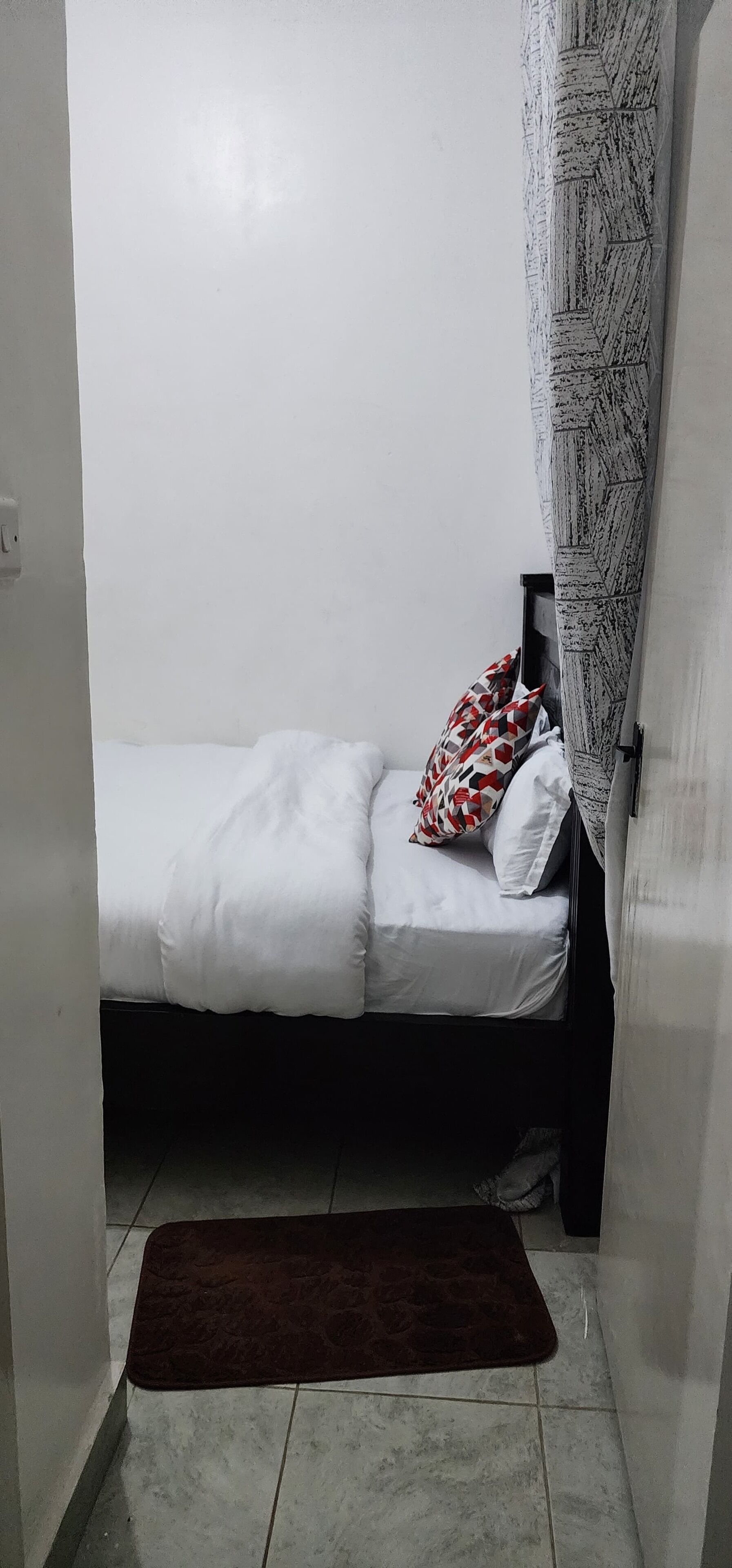 Una habitación pequeña con cama individual, cabecera oscura, ropa de cama blanca y cortina estampada.
