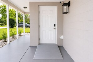 A white door with a security camera above it, a small step leading to the door, and a covered porch area with a light fixture. - 4-bedroom house in perfect Cocoa with cool AC for relaxing stays (Cocoa)