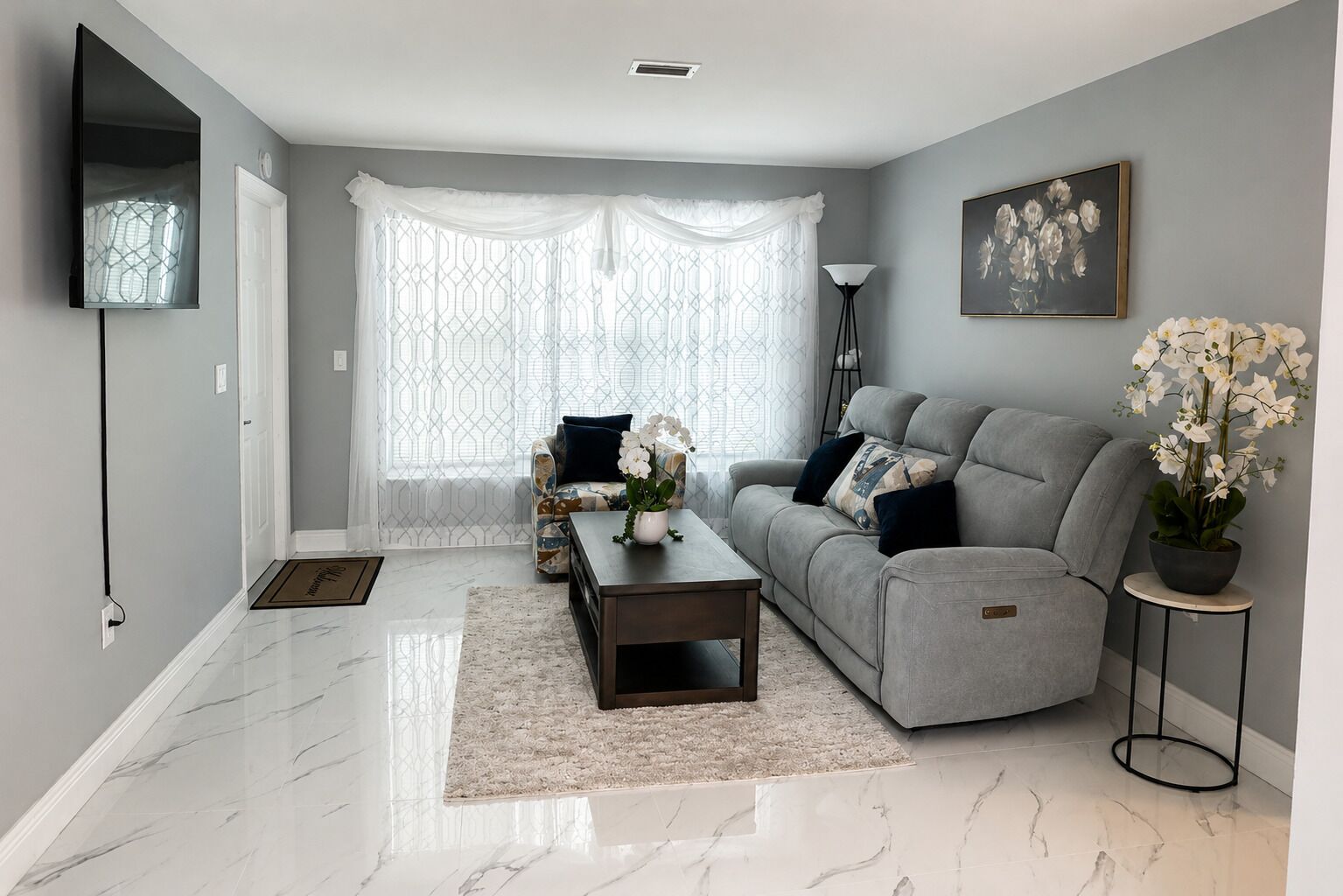 A modern living room with a grey sofa, a coffee table, a patterned armchair, a potted plant, and a framed picture on the wall.