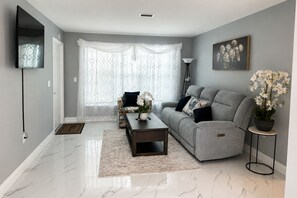 A modern living room with a grey sofa, a coffee table, a patterned armchair, a potted plant, and a framed picture on the wall. - 4-bedroom house in perfect Cocoa with cool AC for relaxing stays (Cocoa)