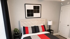 Ein Schlafzimmer mit einem Bett, einem Deckenventilator, einem Nachttisch mit Lampe, einem gerahmten Bild an der Wand und einer Tür.