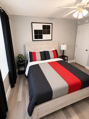 A bedroom with a bed, a ceiling fan, a nightstand with a lamp, a framed picture on the wall, and a door. - 4-bedroom house in perfect Cocoa with cool AC for relaxing stays (Cocoa)