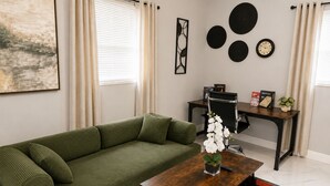 Ein Wohnzimmer mit einem grünen Sofa, einem Holztisch, einem Schreibtisch und einem Stuhl. An den Wänden befinden sich Dekorationen und ein Fenster mit Vorhängen.
