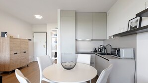A modern kitchen with white cabinets, a microwave, and a kettle. There is a dining area with a round table and white chairs.