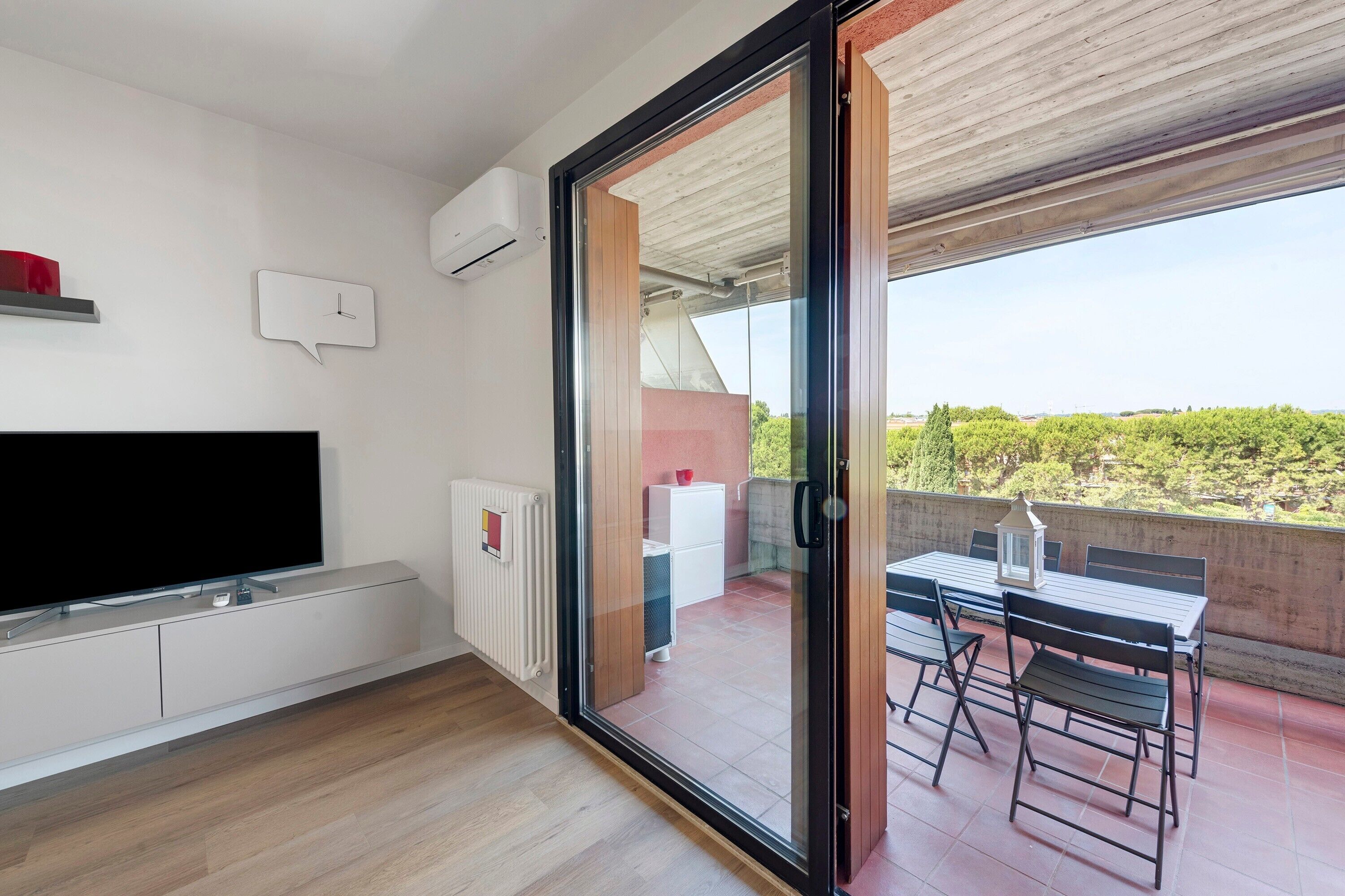 Una sala moderna con un televisor de pantalla plana, un banco empotrado y un ventanal de vidrio que da a un patio exterior con una mesa y sillas.