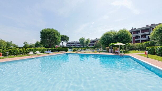 A rectangular swimming pool with clear blue water, surrounded by greenery and a building with multiple floors in the background.