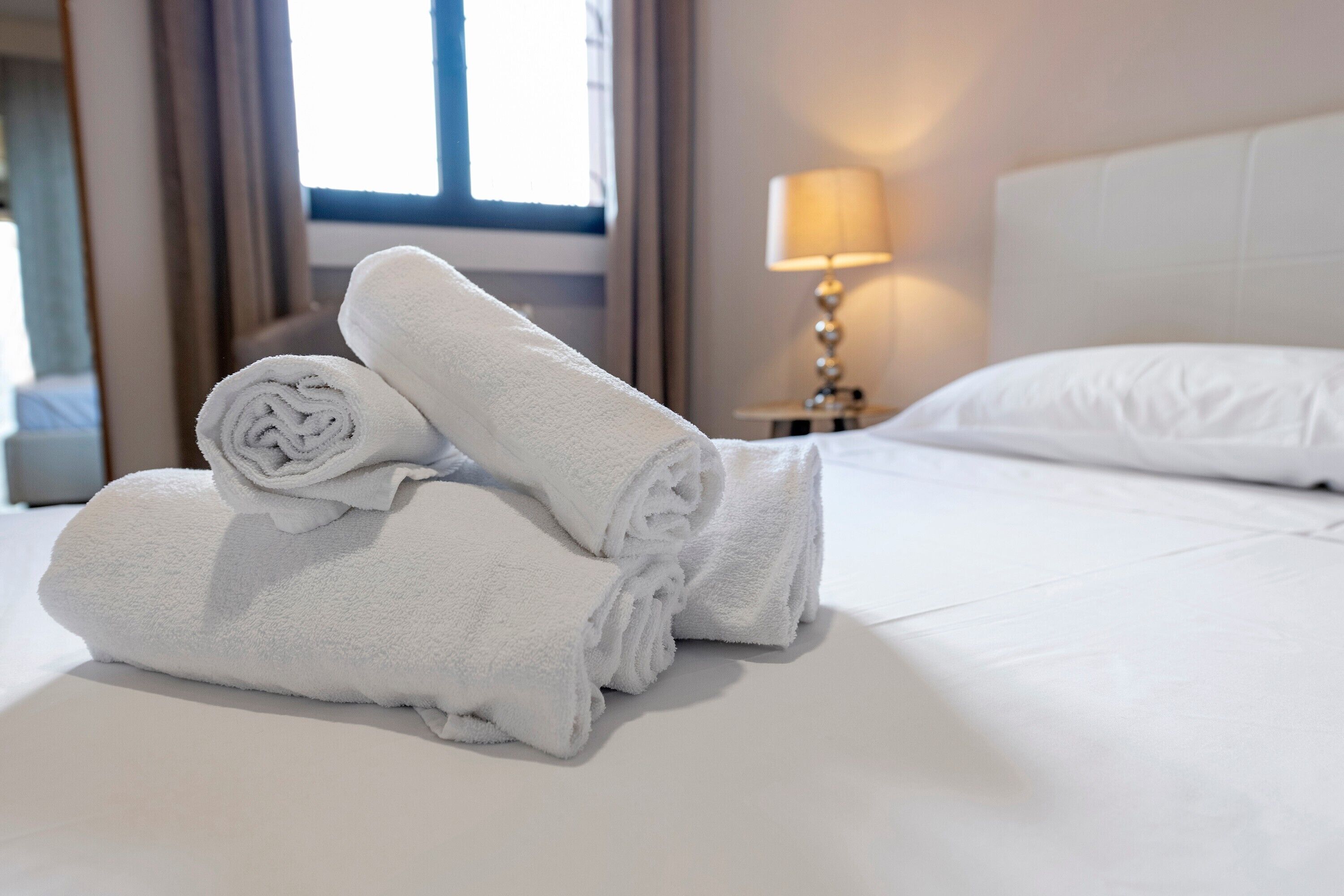 Una cama bien hecha, toallas blancas enrolladas, una lámpara y una ventana al fondo.