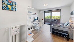 A modern living room with a sofa, a coffee table, and a dining area with a table and chairs. There is a radiator, a wall-mounted shelf with items, and a window with a view.