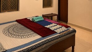A room with a bed, two windows with metal grills, a wooden door, an air conditioner, and a standing fan.