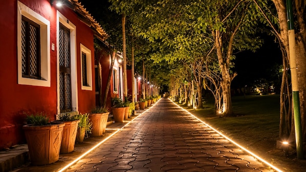 A pathway lined with potted plants and illuminated by a linear light strip, leading to a building with red walls and white window frames.