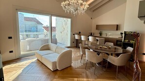 Een ruime woonkamer met een visgraat parketvloer, een witte bank, een eettafel met stoelen, en een kroonluchter.