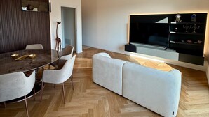 Een moderne woonkamer met een houten visgraatvloer, een glazen eettafel met stoelen, een witte bank, een kroonluchter en een flatscreen-televisie.