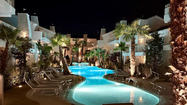 Een zwembadterras van een resort bij nacht, met ligbedden, palmbomen en moderne gebouwen.