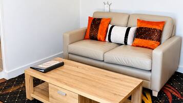 Ein Wohnzimmer mit einer Couch, einem Couchtisch, einem an der Wand befestigten Flachbildfernseher und zwei gerahmten Kunstwerken.