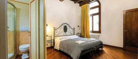 Un dormitori amb sòl de fusta, un llit amb capçalera, una finestra amb cortines, un radiador i una porta.