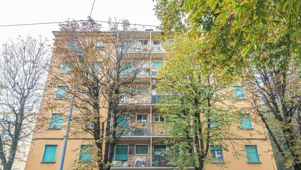 En byggnad med flera våningar, balkonger, en tydligt markerad entré på bottenvåningen och träd framför.