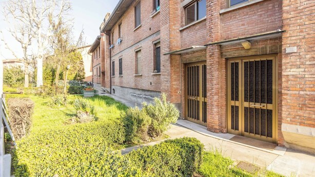 Ein mehrgeschossiges Backsteingebäude mit einem verschlossenen Eingang, ein kleiner Garten mit verschiedenen Pflanzen und einem klaren Himmel.