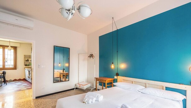 Habitación de hotel con cama, escritorio, silla, espejo y una kitchenette al fondo.
