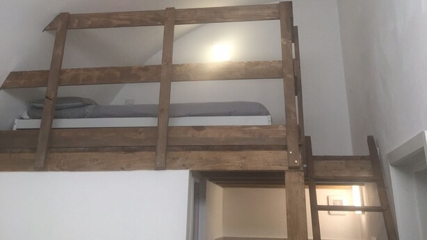 Habitación tipo loft con una cama nido de madera y una escalera, dos bombillas colgantes y techo blanco.