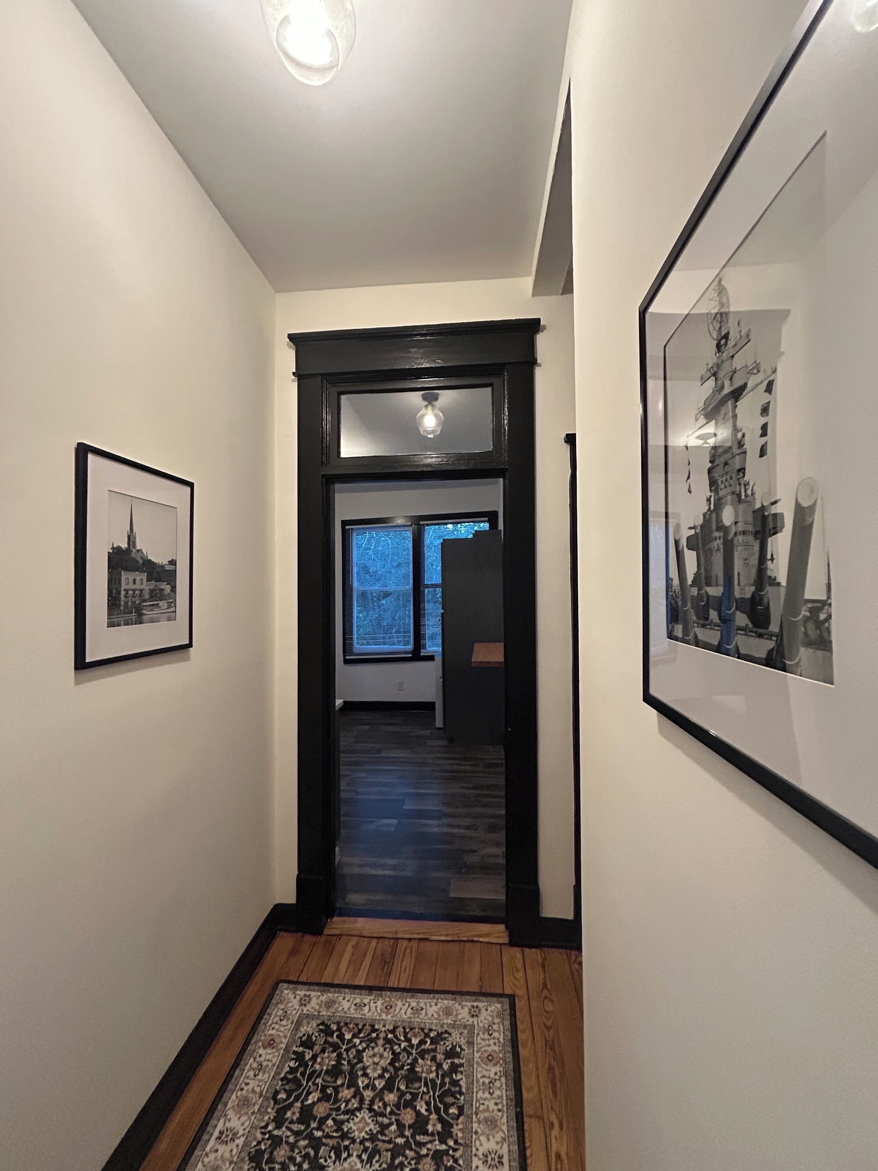 A hallway with framed artwork, a wooden floor, and a door leading to another room.