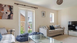 A living room with a sofa, a glass coffee table, a television, and a sliding glass door leading to a balcony. - Spacious Home near Bristol & Bath, Ideal for (Emersons Green)