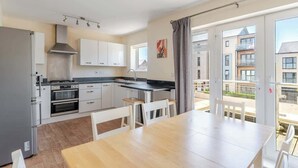 A modern kitchen with stainless steel appliances, a dining area with a wooden table, and a view of residential buildings through large windows. - Spacious Home near Bristol & Bath, Ideal for (Emersons Green)