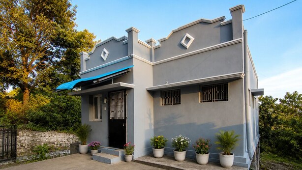 A two-story house with a grey exterior, a blue awning, and a garden with potted plants.
