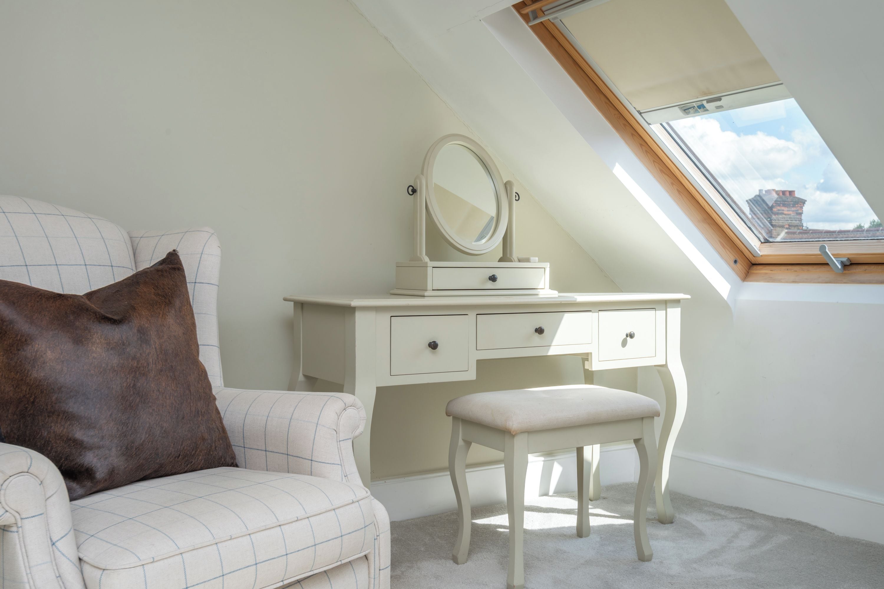 A bedroom with a sloped ceiling, a vanity with a mirror, a chair, and a window with a view of buildings.