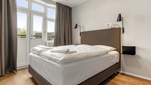 A modern hotel room with a large bed, a bedside table, a wall-mounted lamp, and a window with curtains.