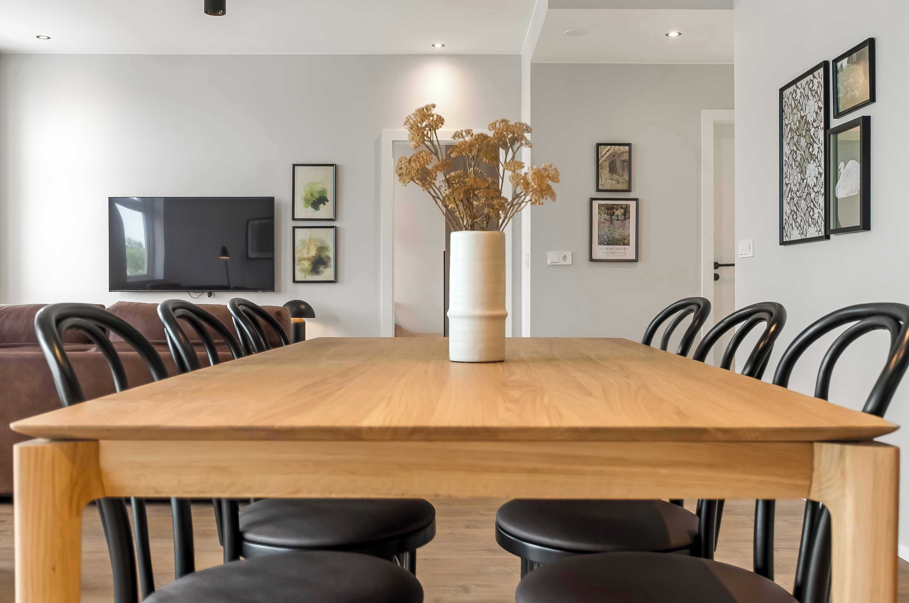 A dining area with a wooden table, black chairs, a vase with dried flowers, and framed artwork on the walls.