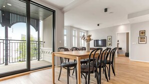 A spacious living area with a wooden dining table, black chairs, a TV, and a balcony with a view.