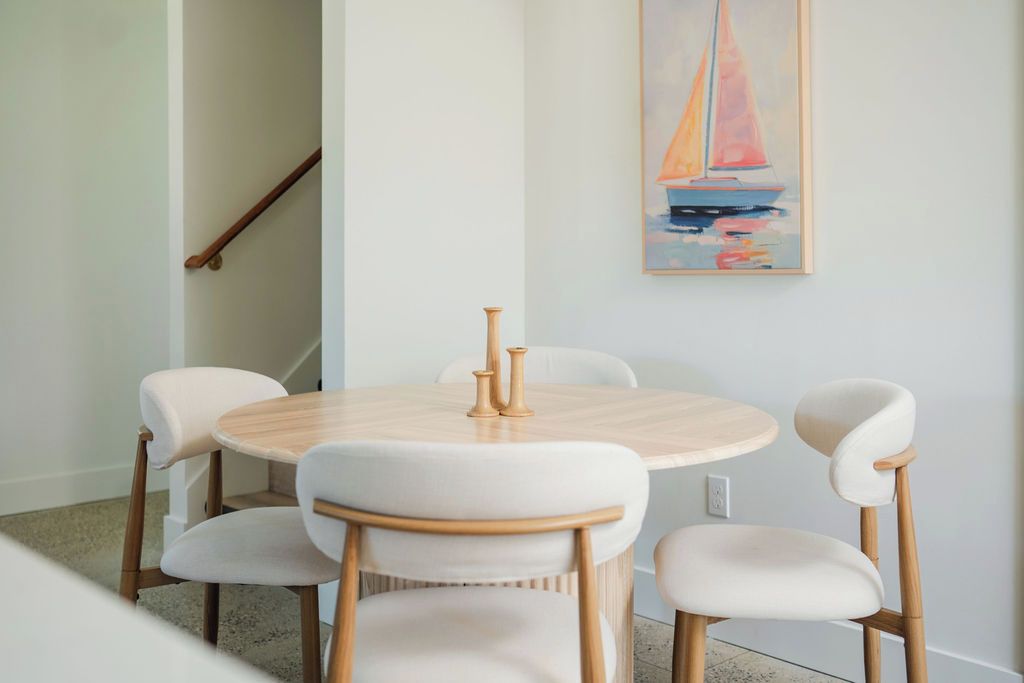 Uma área de refeições com uma mesa redonda e quatro cadeiras, um quadro de um veleiro na parede e uma escada de madeira ao fundo.