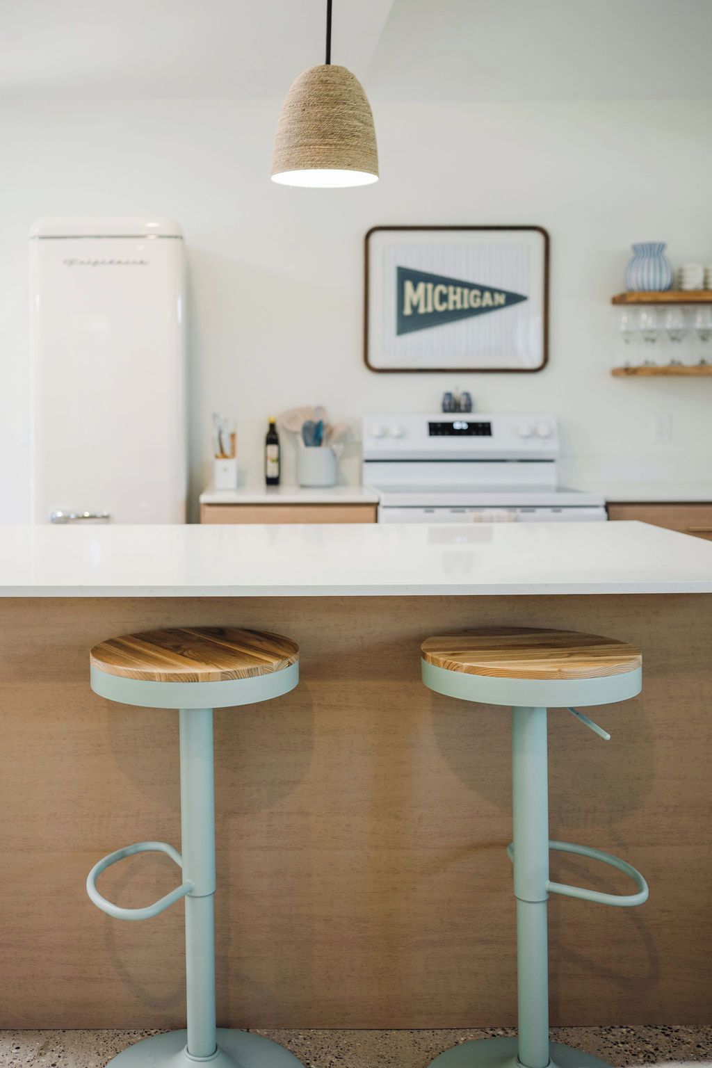 A kitchen with a white countertop, two wooden stools with metal legs, a hanging light fixture, a refrigerator, a stove, and a framed sign on the wall.