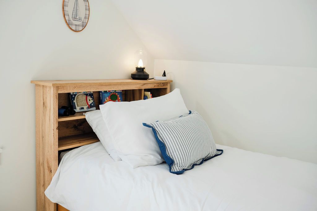 A neatly made bed with white bedding and two pillows, a wooden nightstand with a lamp and books, and a wall-mounted decorative item.