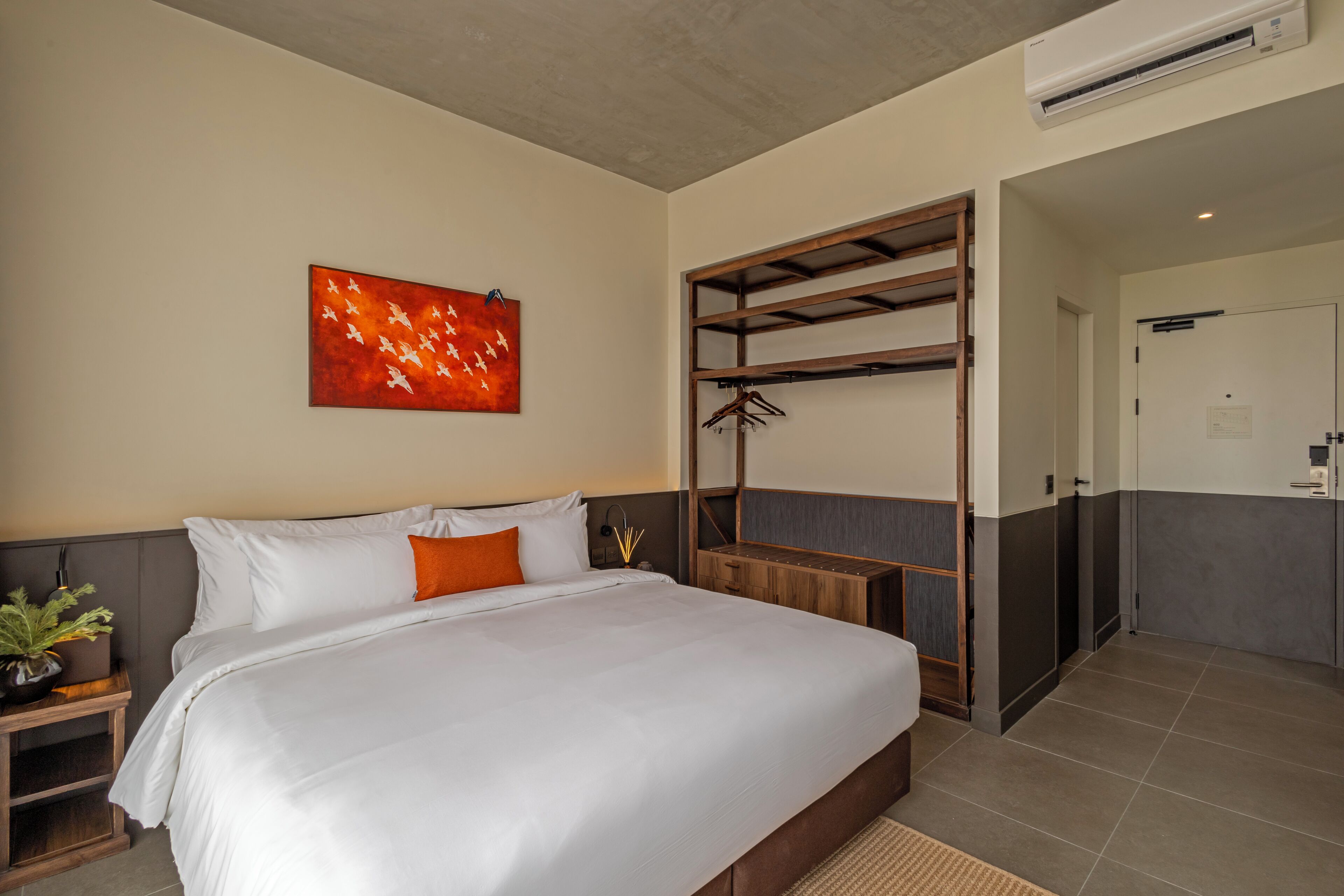 A hotel room with a bed, a wall-mounted artwork, a wooden shelf with clothes, and a door leading to another room.