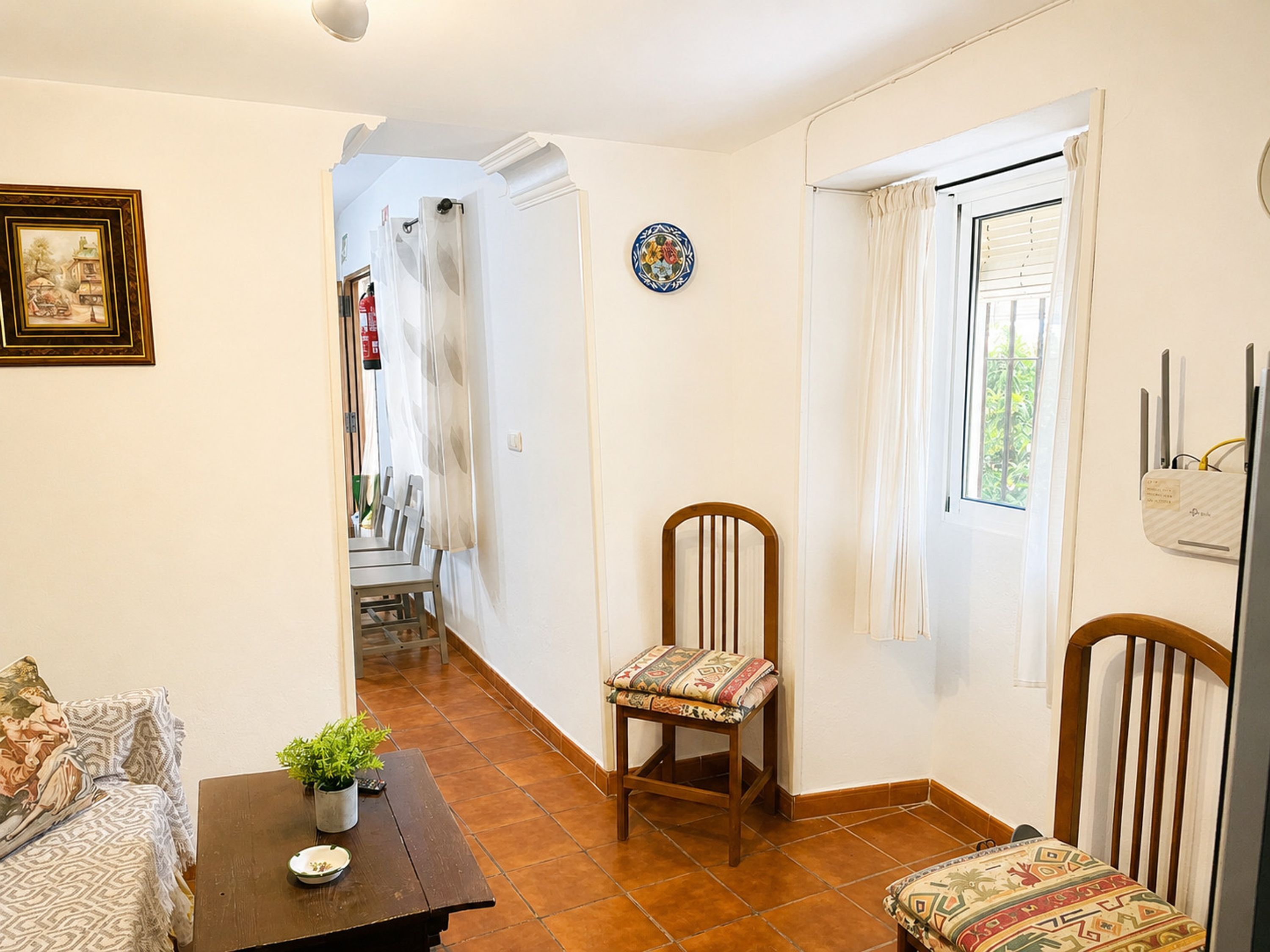 A room with a wooden chair, a small table with a plant, a decorative plate on the wall, and a window with curtains.