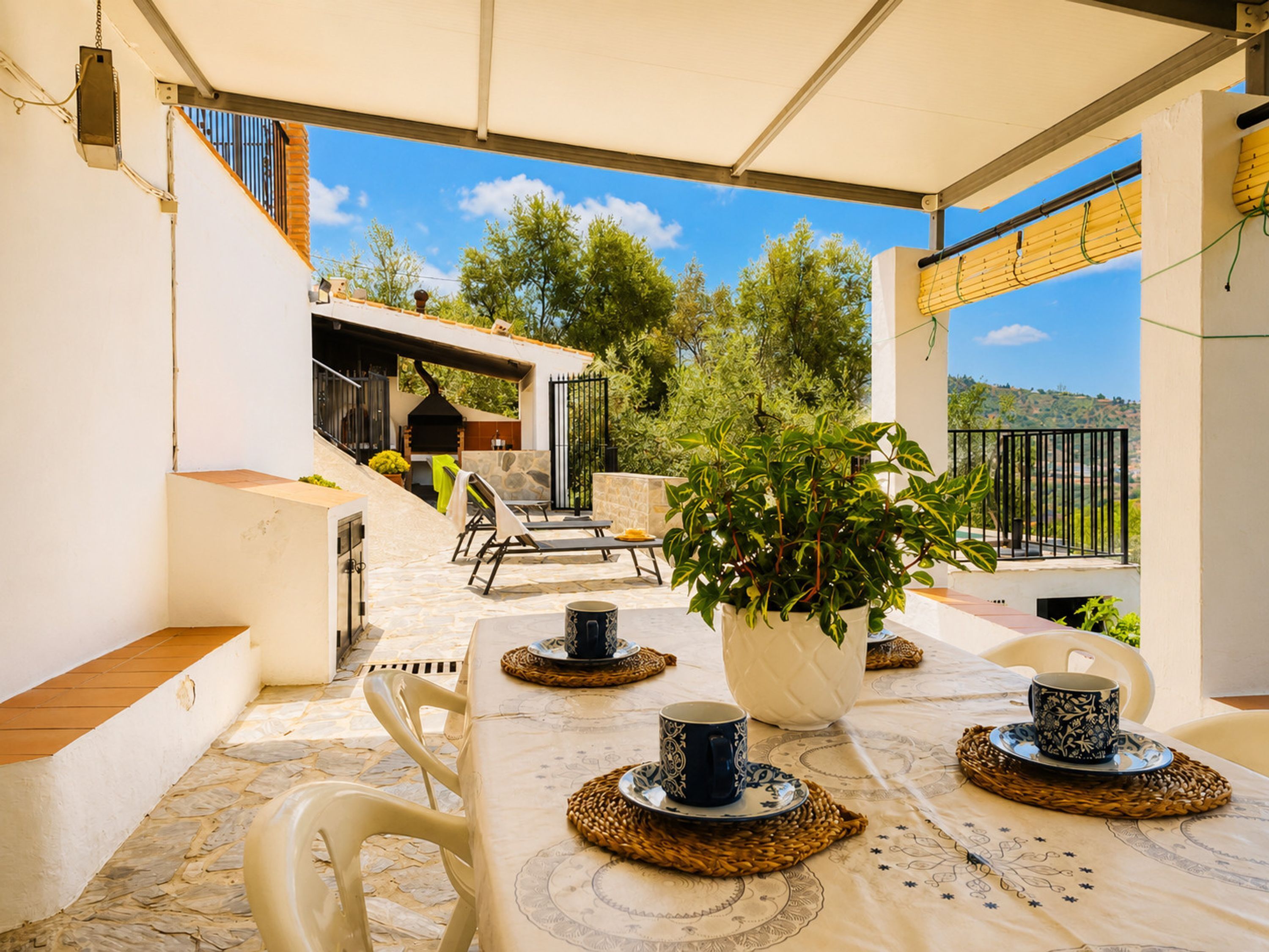 An outdoor patio with a table set for two, a potted plant, and a grill area in the background.