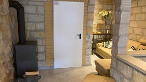 A stone-walled room with a white door, a black heater, a sofa, and a small table with a vase of flowers.
