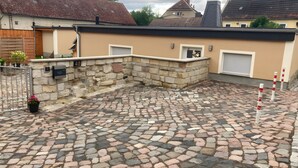 A courtyard with cobblestone flooring, a stone wall, a metal gate, and a building with a beige facade and a garage door.