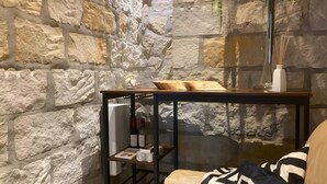 A stone wall interior with a wooden table holding a bowl and a glass vase, a shelf with books and bottles, and a chair with a patterned cushion.