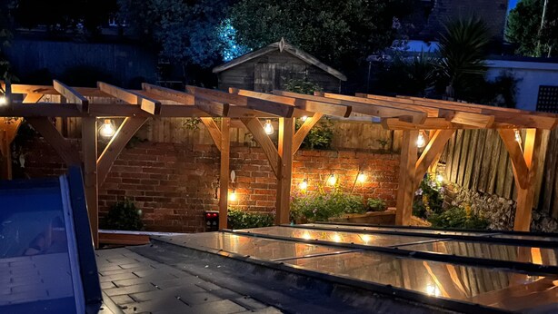 Un espacio al aire libre cubierto, con pared de ladrillo, vigas de madera y plantas en macetas.