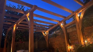 Una estructura de pérgola de madera con pared de ladrillo, un banco y luces colgantes.