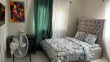 A bedroom with a bed, a standing fan, a desk, and a painting on the wall.