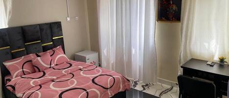 A bedroom with a bed, a desk, a chair, a white air conditioner, and a painting on the wall.