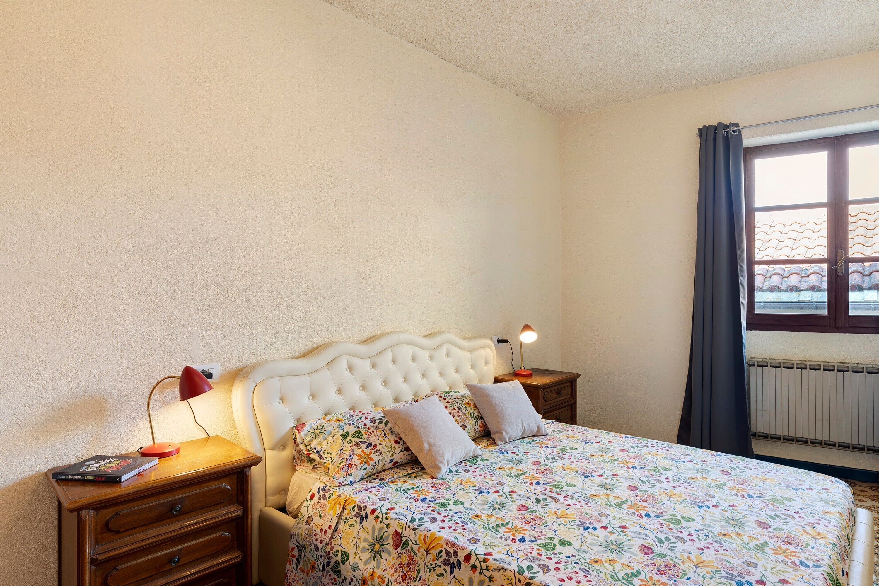 Une chambre à coucher avec un lit, une table de chevet, une lampe, une fenêtre avec des rideaux et un radiateur.