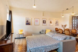 A hotel room with a bed, a desk with a lamp, a television, and framed pictures on the wall.